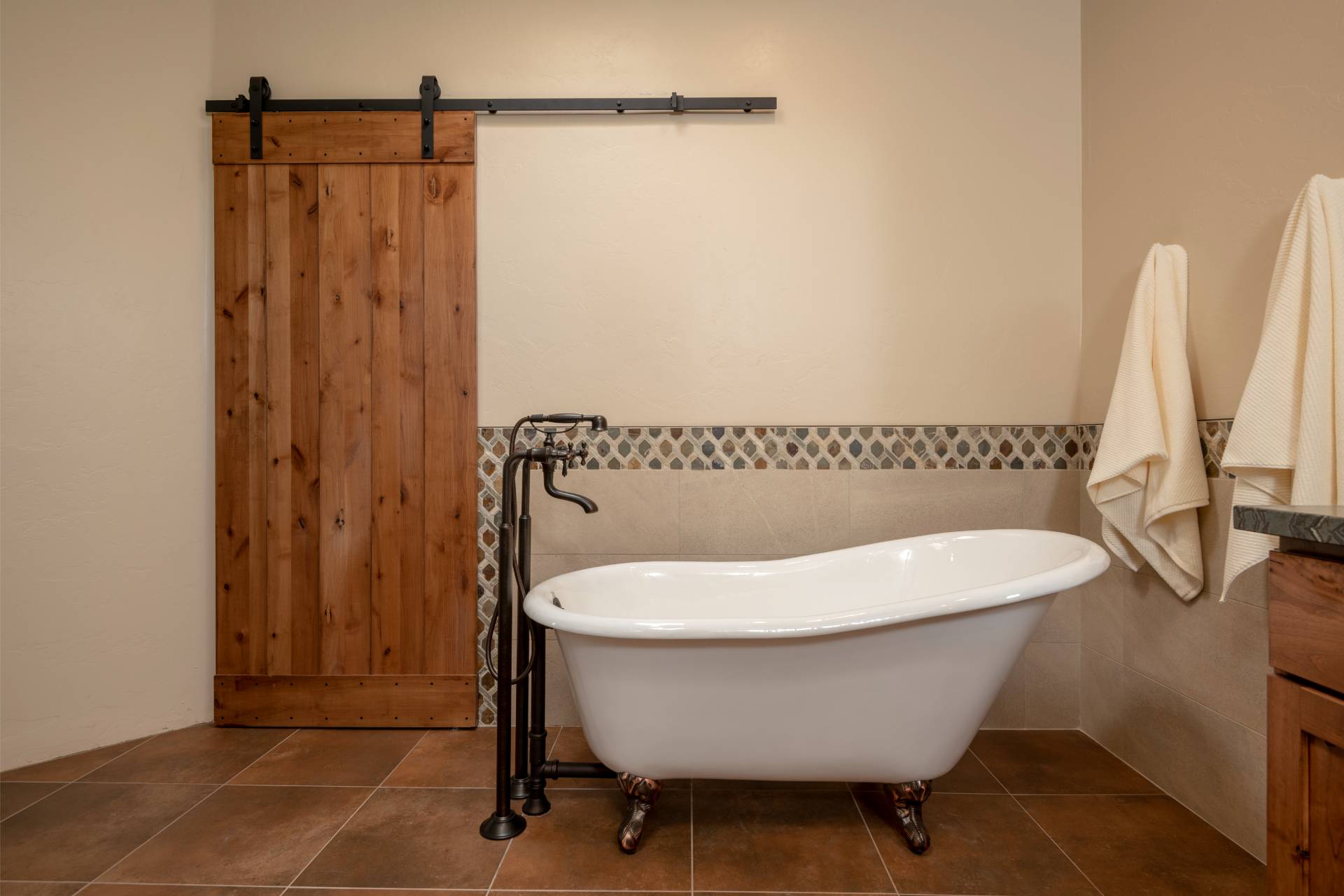 White cast-iron clawfoot slipper tub with an oil-rubbed-bronze floor filler, a multicolor slate mosaic tile band, and a knotty-alder sliding barn door