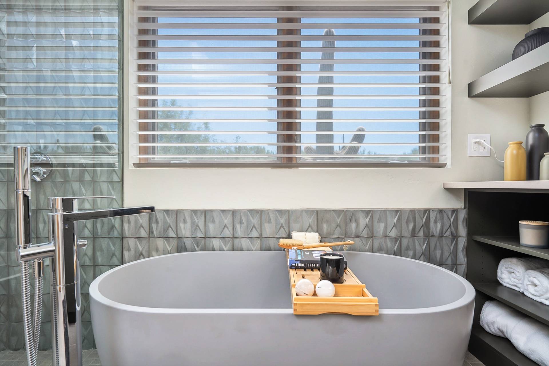 Garden Drive soaking tub set below a wide window framing a saguaro, with dimensional gray tile and open shelving