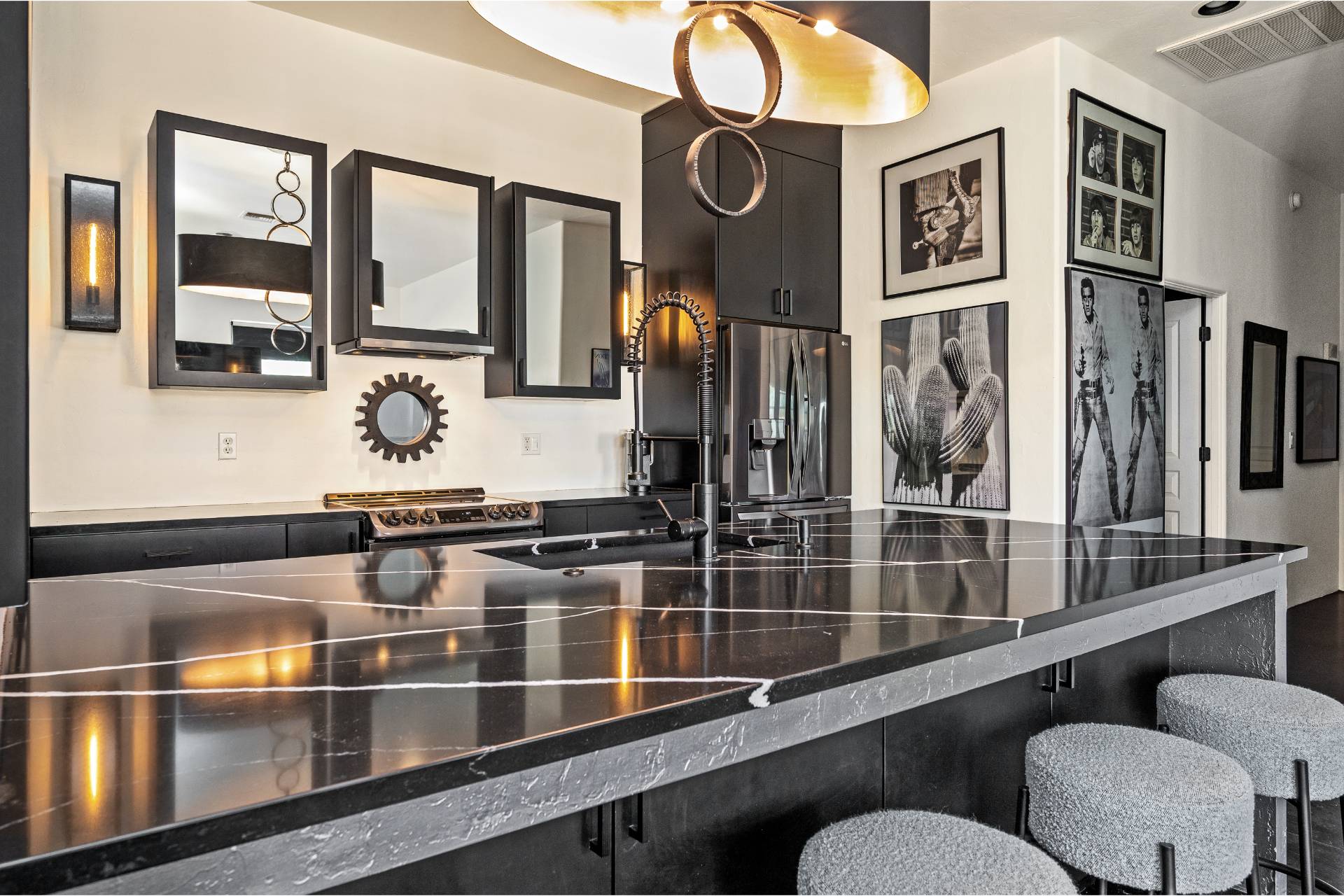 Low-angle view across the Gassel marble island toward the range wall, black cabinet-front refrigerator, gear-form mirror, and framed pop-art prints