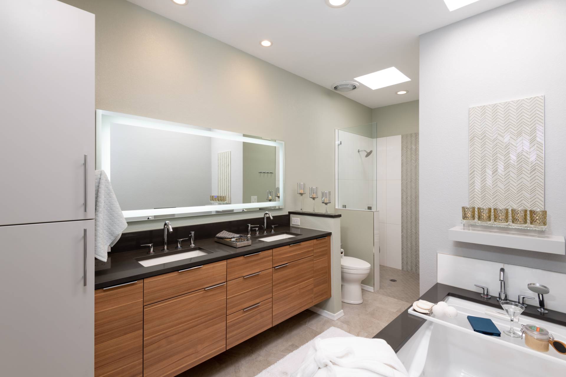 greenberg-primary-bathroom-01 Greenberg primary bath with a floating walnut double vanity, honed black-stone counter, backlit rectangular mirror, curbless glass shower, and a drop-in soaking tub