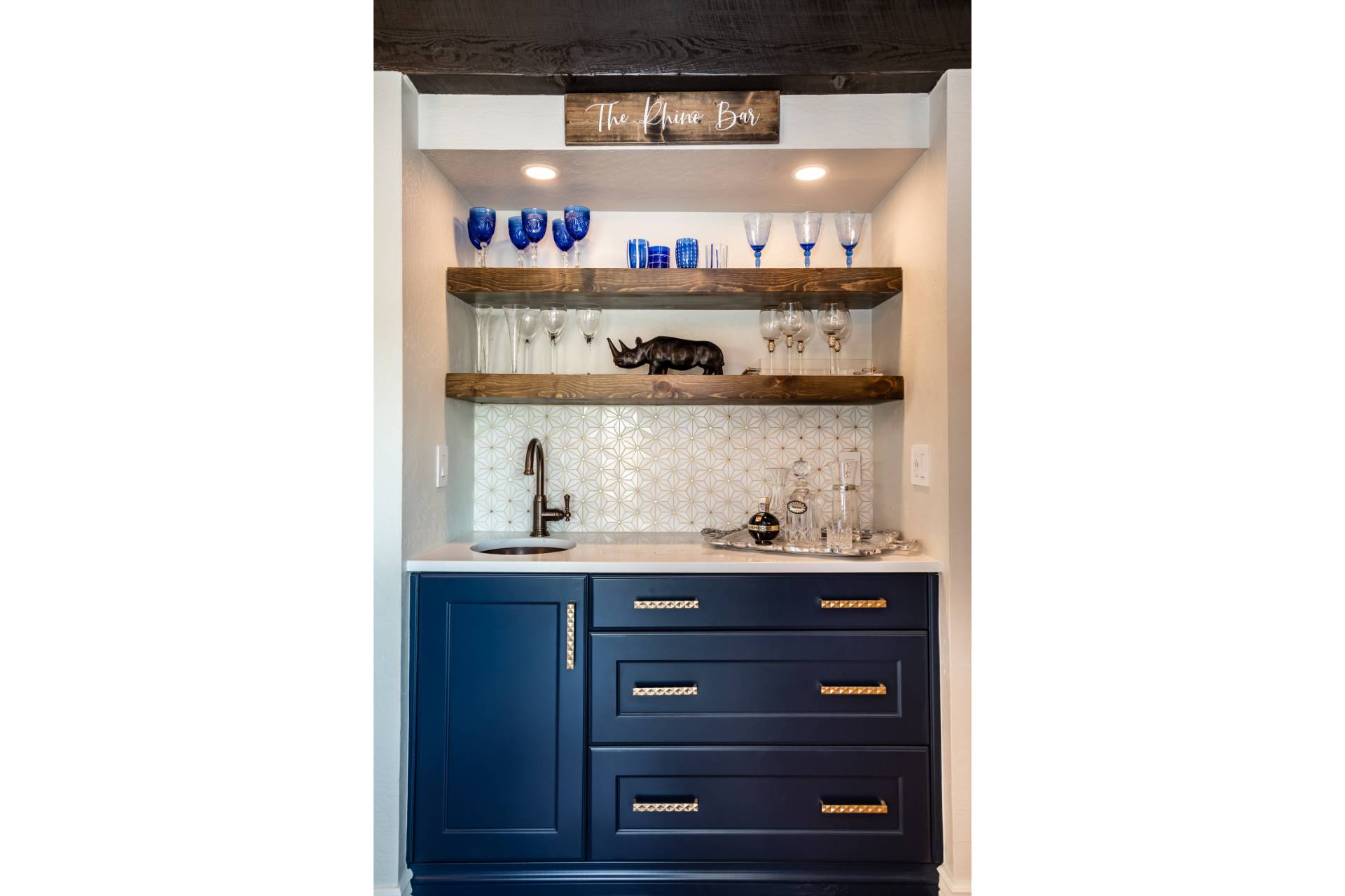 Keeley built-in bar station in navy cabinetry with a star-patterned tile backsplash, stained-wood floating shelves, and the “Rhino Bar” sign