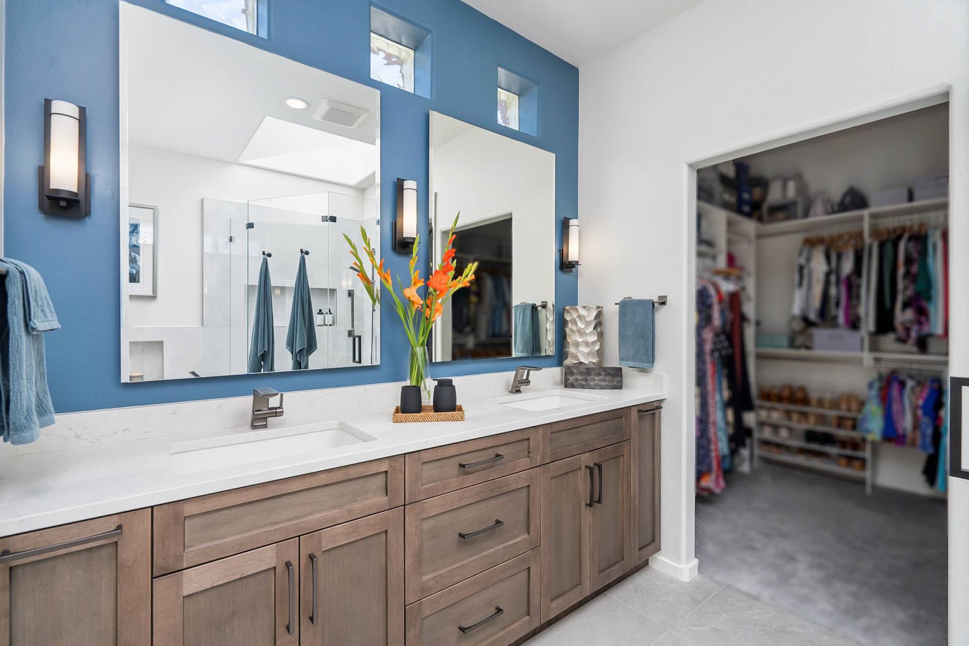 Lucente double vanity in stained wood with a quartz top, twin mirrors on a blue accent wall, and the adjoining walk-in closet