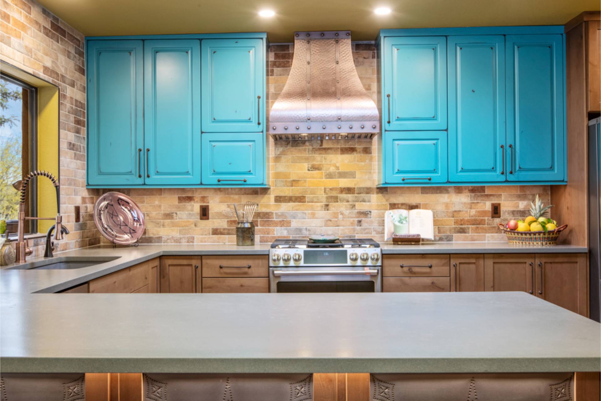 Close view of the Vaughn range wall: hammered-copper hood centered between turquoise cabinets above a stainless gas range and travertine brick backsplash